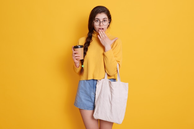 Female holding reusable coffee mug and cotton bag while posing isolated over yellow wall, having astonished facial expression, covering her widely opened mouth with palm.