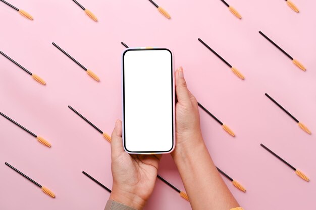 Female holding smart phone with eyelashes brush on pink background