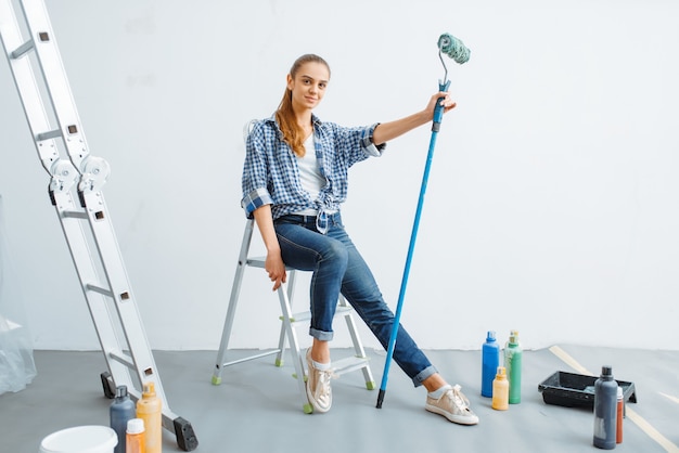 Female house painter with paint roller sitting on a ladder. Home repair, happy woman doing appartment renovation