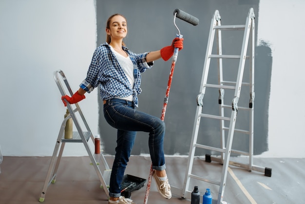 Female house painter with paint roller sitting on a ladder. Home repair, happy woman doing appartment renovation