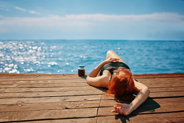 写真 スイムスーツを着た女性がレモネードのボトルと赤毛で日光浴をしている木製の埠頭に横たわっています ⁇