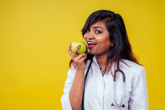 Femmina indiana giovane e bella donna bionda ginecologo medico utilizzando uno stetoscopio tenendo una mela verde in mano in un camice medico bianco su sfondo giallo in studio.