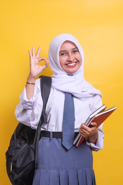 Female Indonesian high school muslim student in white and grey uniform holding books while giving ok