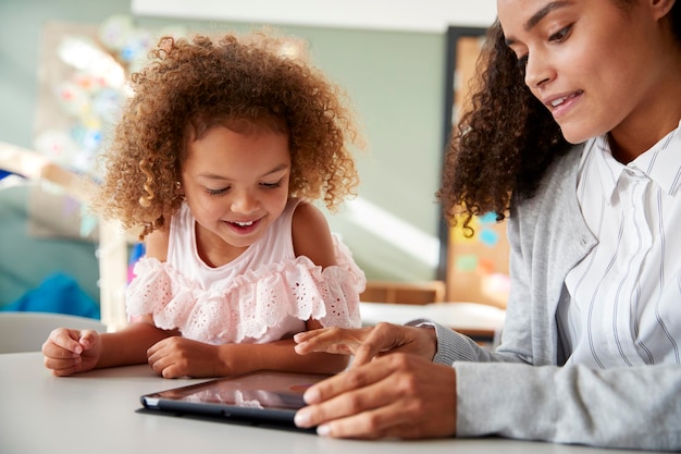 Female infant school teacher using a tablet computer working one on one in a classroom with a young mixed race schoolgirl selective focus close up