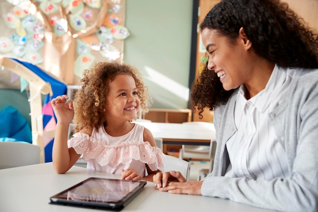 Female infant school teacher working one on one in a classroom using a tablet computer with a young mixed race schoolgirl smiling at each other close up