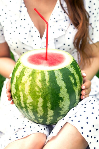 Female is holding watermelon with cocktail straw on background of her white dress.