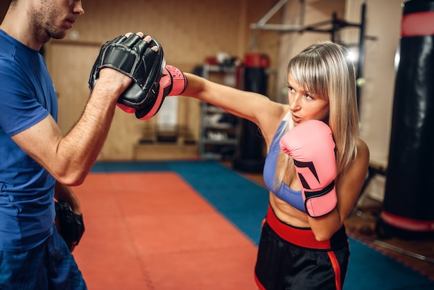 Female kickboxer on workout with male personal trainer in pads, gym interior. Woman boxer makes hand punch on training, kickboxing practice