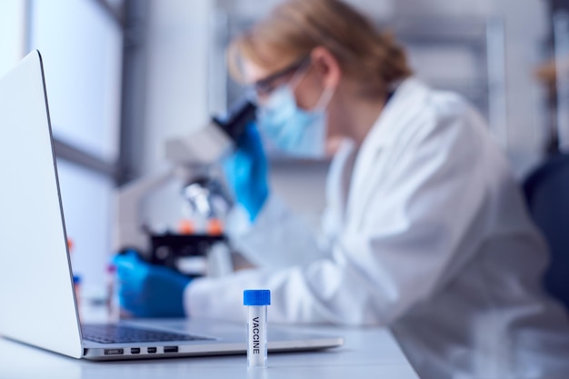 Female lab worker wearing lab coat working on vaccine in
laboratory with laptop and microscope