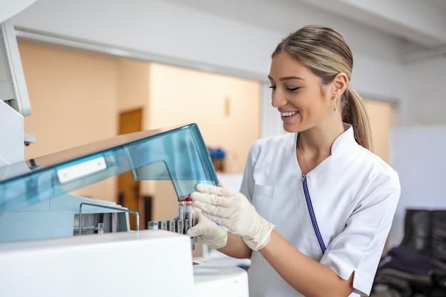 Foto assistente di laboratorio femminile che effettua analisi con provette e macchine analizzatrici sedute presso il moderno laboratorio tecnico che carica campioni in un analizzatore chimico