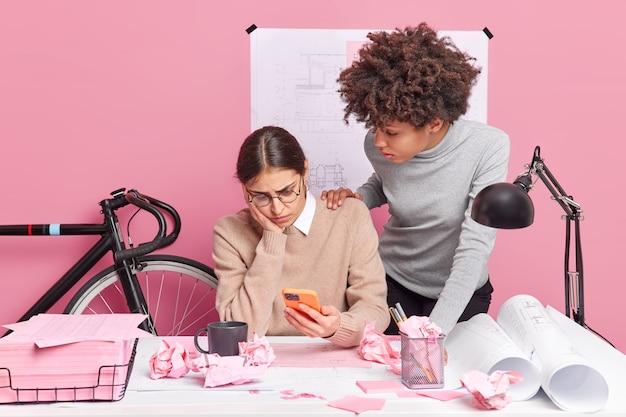 Female leader of architectural project failed to make blueprints of building holds smartphone concentrated with puzzled expression at display her Afro American partner tries to help gives advice