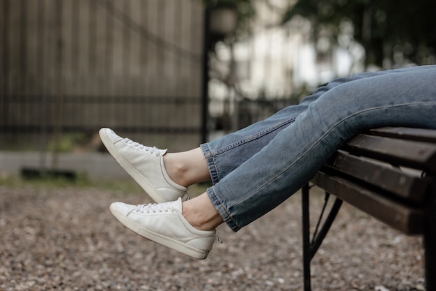 Photo female legs in comfortable casual sneakers sitting on the bench in the park. sneakers mock-up.