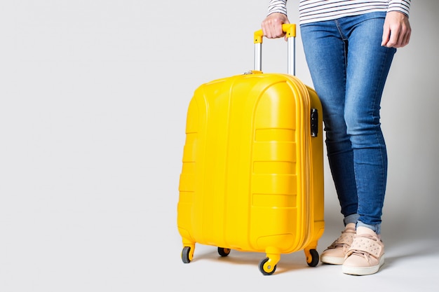 Female legs in jeans stand next to a yellow suitcase on a light background