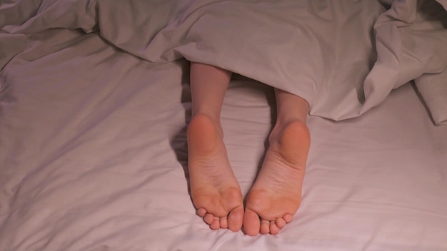 female legs joyfully movie and dance with toes while lying in bed on a sheet under blanket Happy woman girl with bare legs foot is relaxing and rest on a bed in a home bedroom in the morning