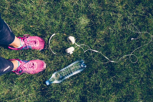 Female legs in pink sneakers, white headphones and a plastic bottle of water on the grass