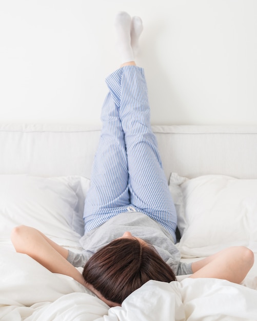 Female legs raised up high and arms under her head lying on bed in bedroom wearing pajamas