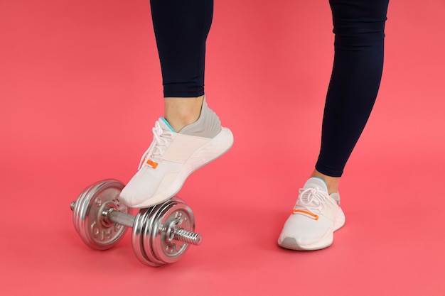 Female legs in sportswear and dumbbell on pink background