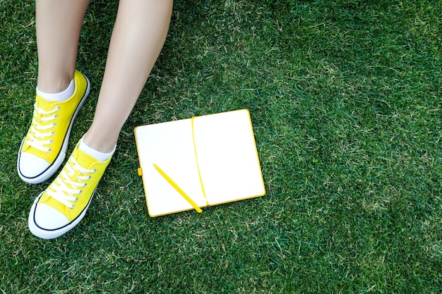 Female legs in yellow sneakers and notebook with pen on grass