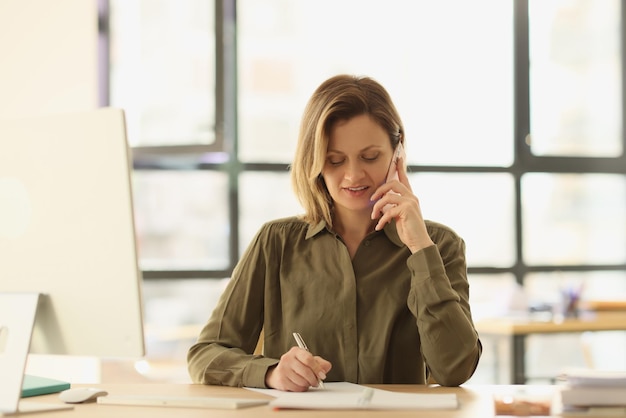 Female manager is talking on phone and taking notes in notebook while sitting at her desk in