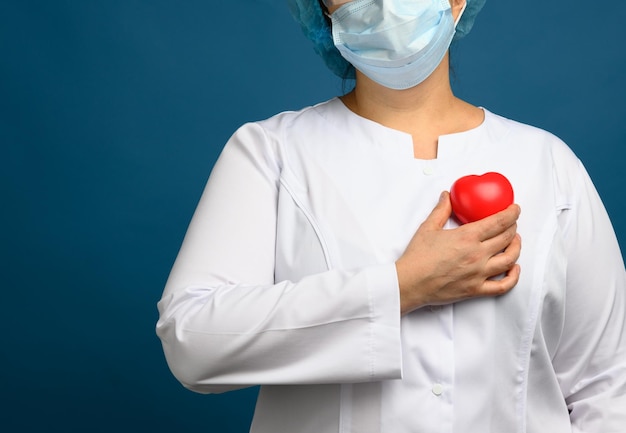 Photo female medic in a white coat a mask stands and holds a red heart on a blue background the con