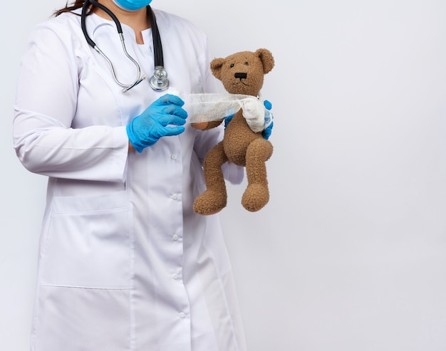 Female medic in a white coat with buttons holding a brown teddy bear and bandaging her paw