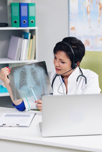 Female medical doctor wears white coat, headset video calling distant patient on laptop