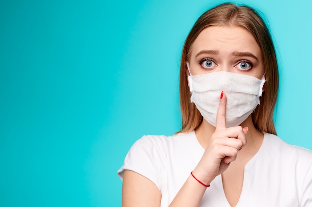 Female in medical mask holding finger near lips on isolated blue