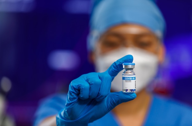 Female medical people wearing a hygiene face mask and protective glass staying in laboratory room, holding Covid-19 vaccine bottle with lab equipment in blur background.