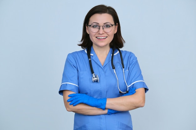 Operaio medico femminile in uniforme blu con stetoscopio e guanti, donna professionale fiduciosa con le braccia conserte che guarda l'obbiettivo, sfondo grigio chiaro