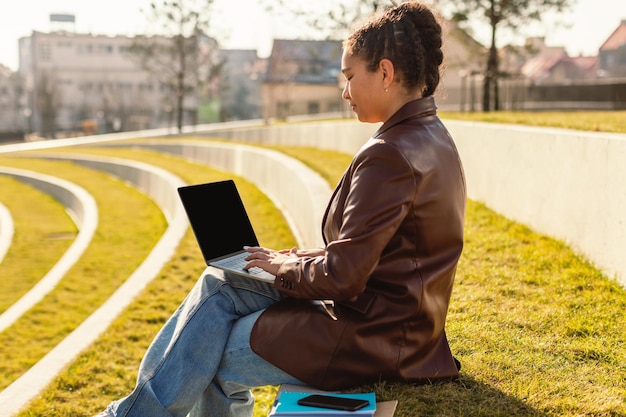 Female mixed race student using laptop with blank screen outdoors sitting in park mockup for