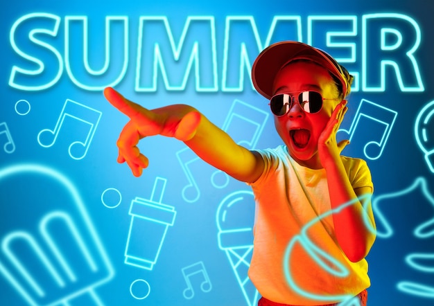 Female model in trendy neon light on blue background. Modern design. Beautiful little girl posing in cap and sunglasses, looks astonished. Concept of facial expression, summer, vacation, music.