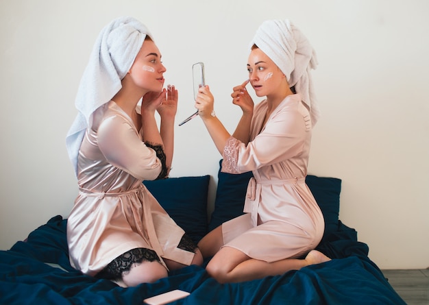 Female models apply cream on face. Two young women in towels and pajamas have a fun spa party together in home.