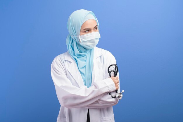 A female muslim doctor with hijab wearing a surgical mask over\
blue background studio.