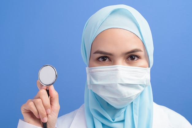 A female muslim doctor with hijab wearing a surgical mask over blue wall