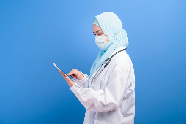 Un medico musulmano femminile con l'hijab che indossa una mascherina chirurgica facendo uso della compressa sopra l'azzurro