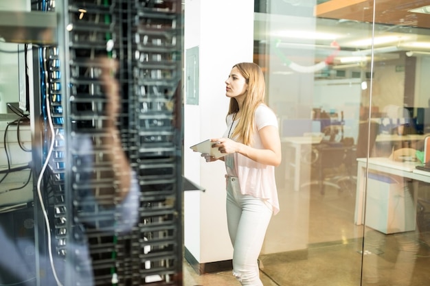 Female network administrator with digital tablet standing in\
data center room and looking at networking device in rack