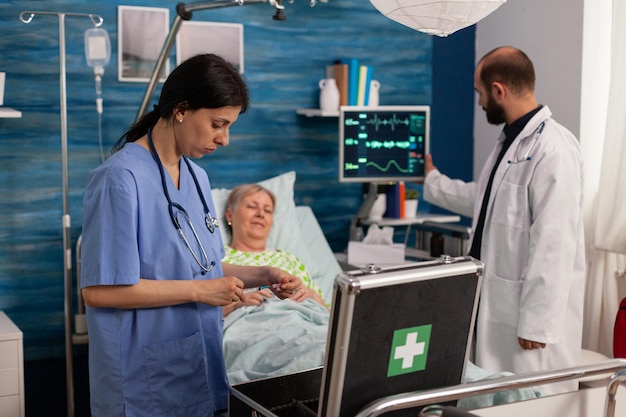 Female nurse preparing medicine with syringe to apply to sick\
elderly woman in hospital room bed. male doctor analyzing vital\
sign values displayed on monitor, professional health care.