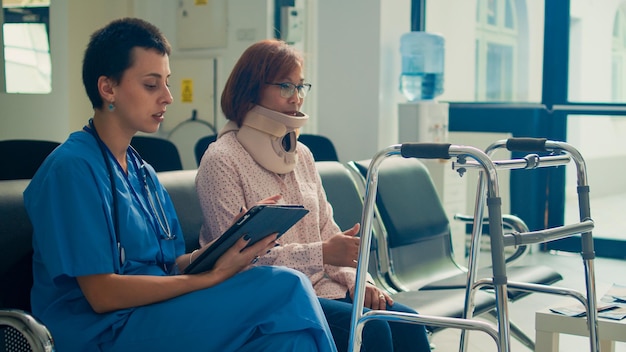 Female nurse talking to asian patient wearing cervical neck collar, taking notes on tablet at medical consultation. Doing examination with injured person, injury pain. Handheld shot.