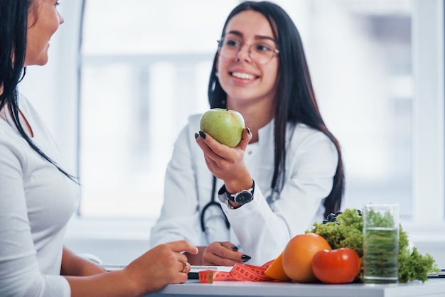 Female nutritionist holding green apple and gives consultation to patient indoors in the office.