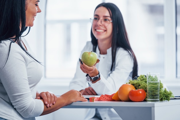 Female nutritionist holding green apple and gives consultation to patient indoors in the office.