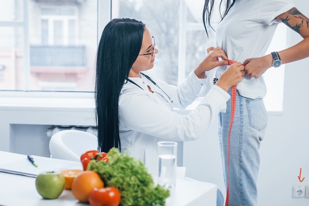 Female nutritionist measuring waist of patient by tape indoors in the office.