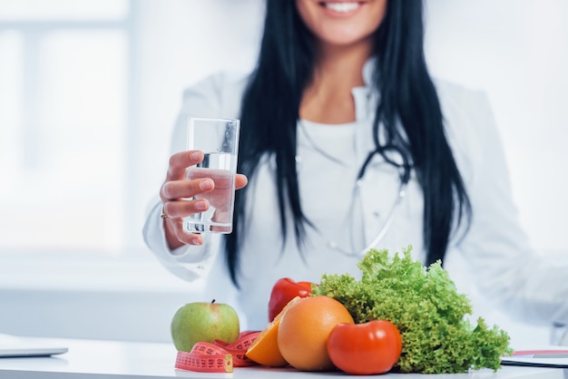 Female nutritionist in white coat sitting indoors in the office at workplace with glass of drink.