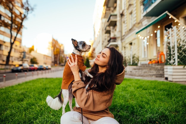 female owner sitting on a green lawn and holding a small cute dog in her arms on street background