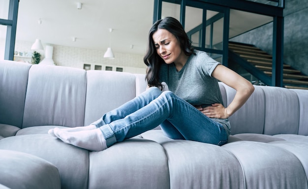 Female pain. Woman with pain in stomach is sitting on couch at home