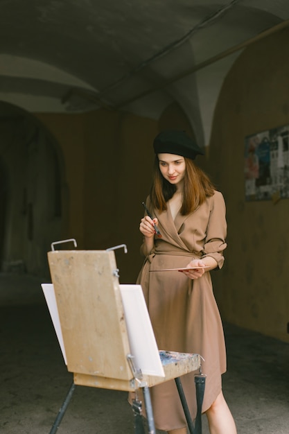 Female painter painting in the city. A woman wearing khaki dress and a black hat