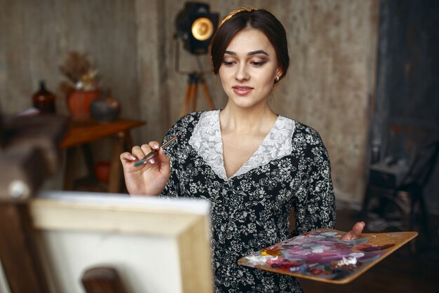 Female painter with brush and palette draws in art studio. Artist at her workplace, creative master at the easel in workshop