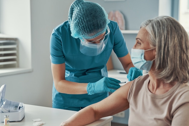 Фельдшер в защитной спецодежде делает прививку от коронавируса зрелой женщине во время работы в больнице