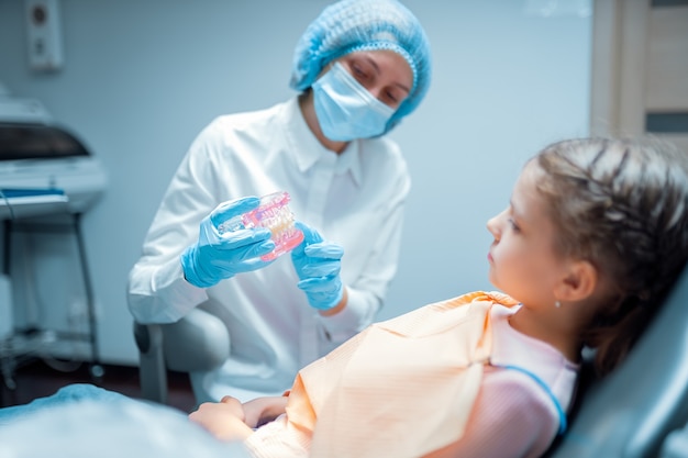 Female pediatric dentist orthodontist teaching little girl patient using teeth model with brecets