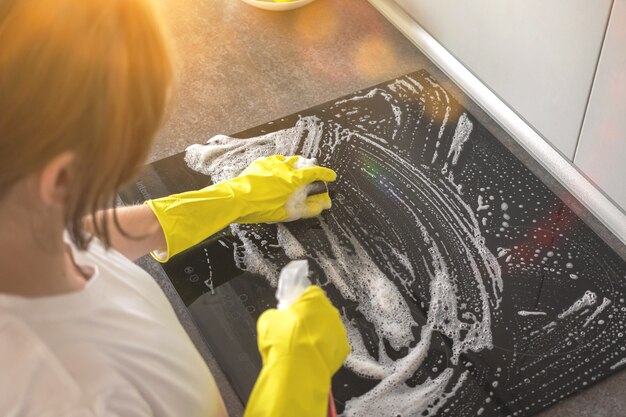 Persona di sesso femminile che pulisce il fornello elettrico con spugna gialla e flacone spray con detergente in cucina, foto dall'alto vista