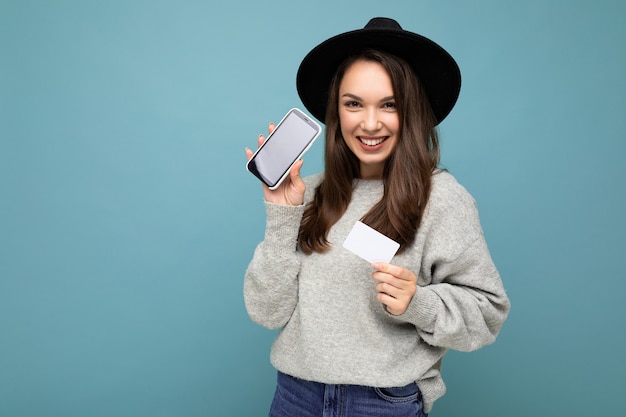 female person wearing black hat and grey sweater isolated over blue wall holding credit card and mobile phone with empty display  .