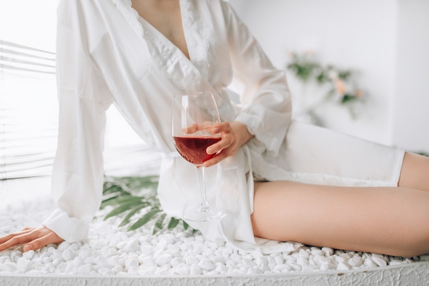 Female person in white bathrobe sitting on the edge of the bath with glass of red wine. Bathroom interior with window